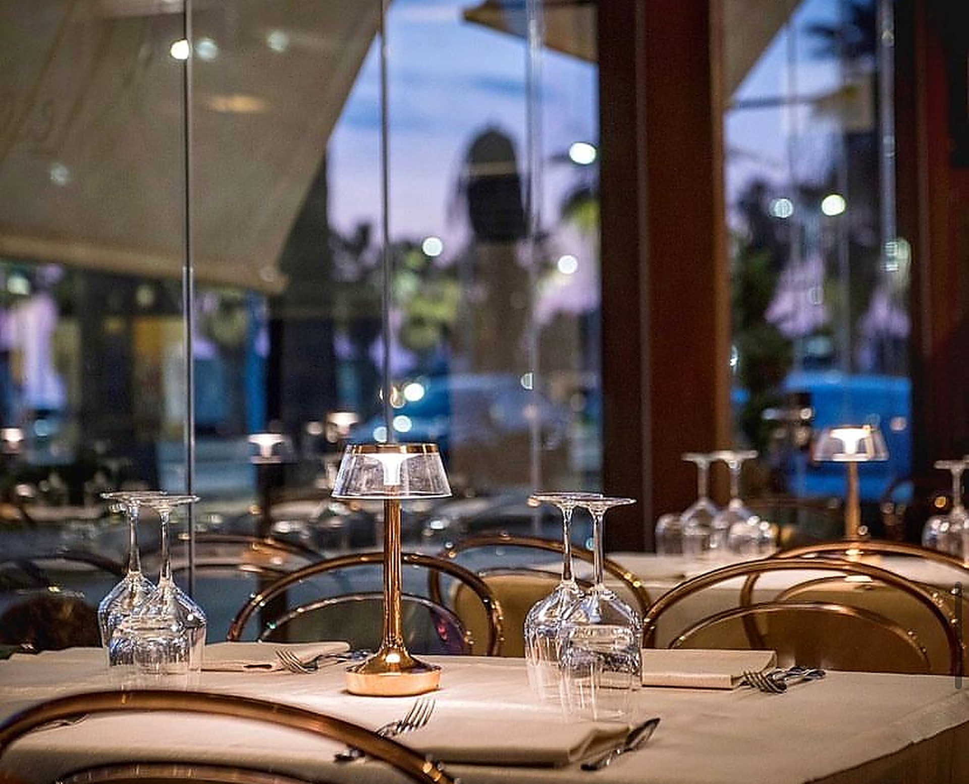Tavolo apparecchiato in trattoria all'imbrunire, calici e lume in ottone, con San Leone che si intravede oltre le vetrate