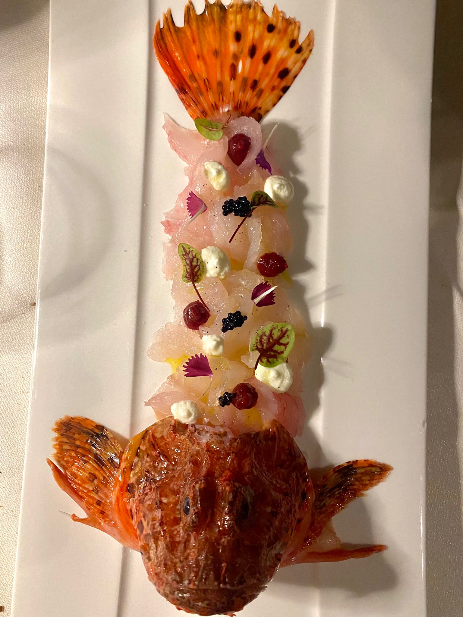 Scorfano intero elaborato dallo chef, con crudo, caviale e fiori eduli disposti tra la testa e la coda del pesce